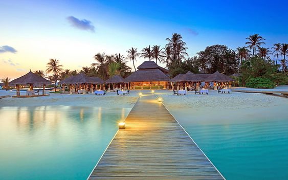 A resort infinity pool at dusk with thatched pavilions along the deck