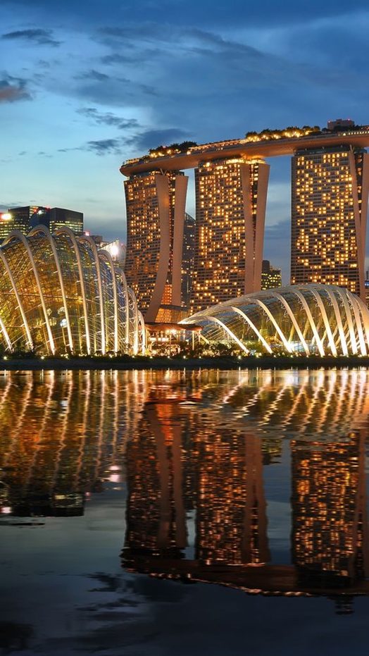 A three-towered hotel with a rooftop deck reflected in the bay at night