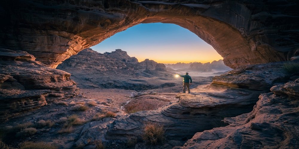 A natural rock arch over a canyon trail with a walker below